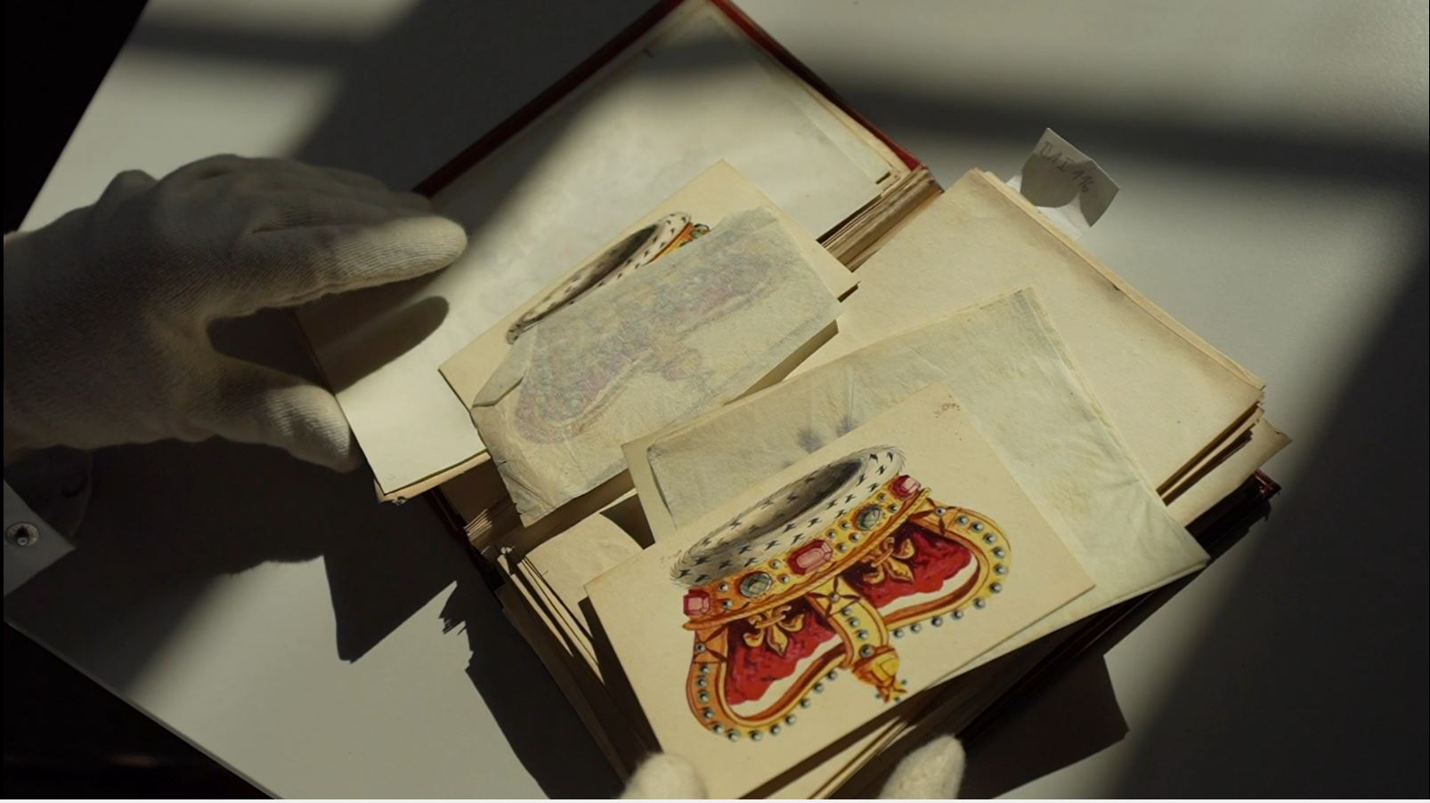 Das Bild zeigt eine Sammlung loser Blätter mit Zeichnungen in einer Mappe und die behandschuhten Hände einer untersuchenden Person.